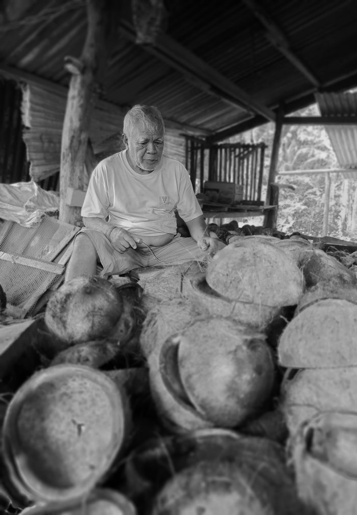 About Elderly man engaged in coconut processing in a rustic, monochrome setting, evoking a sense of traditional work.